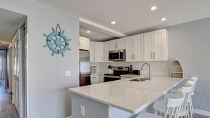 A modern kitchen with white cabinets, a central island with bar stools, a stainless steel microwave, and a ship's wheel decoration. - Sunskipper E2 - 2 bedroom ocean view condo (Carolina Beach)