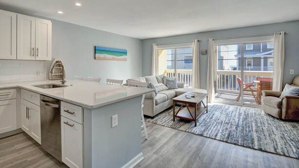 A modern kitchen with white cabinets, a stainless steel sink, and a built-in dishwasher. There is a dining area with a white table and chairs, and a living room with a beige sofa and a coffee table. - Sunskipper E2 - 2 bedroom ocean view condo (Carolina Beach)