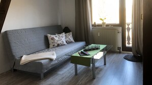 Una sala moderna con un sofá gris, dos cojines estampados, una mesa de centro de vidrio con un mantel verde y una pequeña planta en maceta en el alféizar.