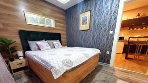 A bedroom with a wooden bed, a nightstand, a large window, and a view of the kitchen.