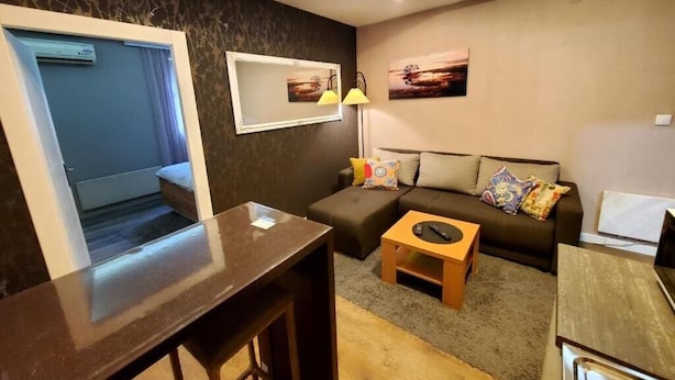 A modern living room with a sofa, a coffee table, and a TV stand. There is a window with curtains, a lamp, and artwork on the wall.