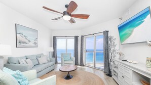Ein modernes Wohnzimmer mit Sofa, Sessel und Couchtisch, einem Deckenventilator, einem Gemälde und einem großen Fenster mit Meerblick.