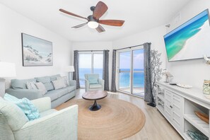 A modern living room with a sofa, armchair, and coffee table, a ceiling fan, a painting, and a large window with a view of the sea. - Renovated 2B Gulf Front Condo @ Emerald Isle 805! (Pensacola Beach)