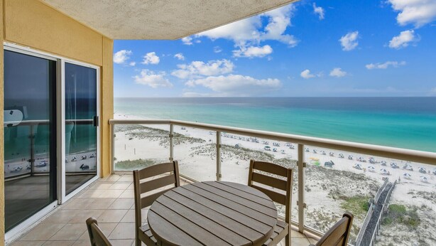 Ein Balkon mit Tisch und Stühlen, der Blick auf einen Strand mit klarem türkisblauem Wasser und einem sandigen Ufer bietet.