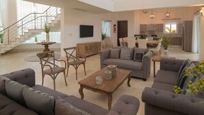 A spacious living area with a sofa set, wooden coffee table, and a dining table. A staircase leads to an upper level, and a kitchen is visible in the background. - Luxury Villas - Mandatory All-Inclusive (Puerto Plata)