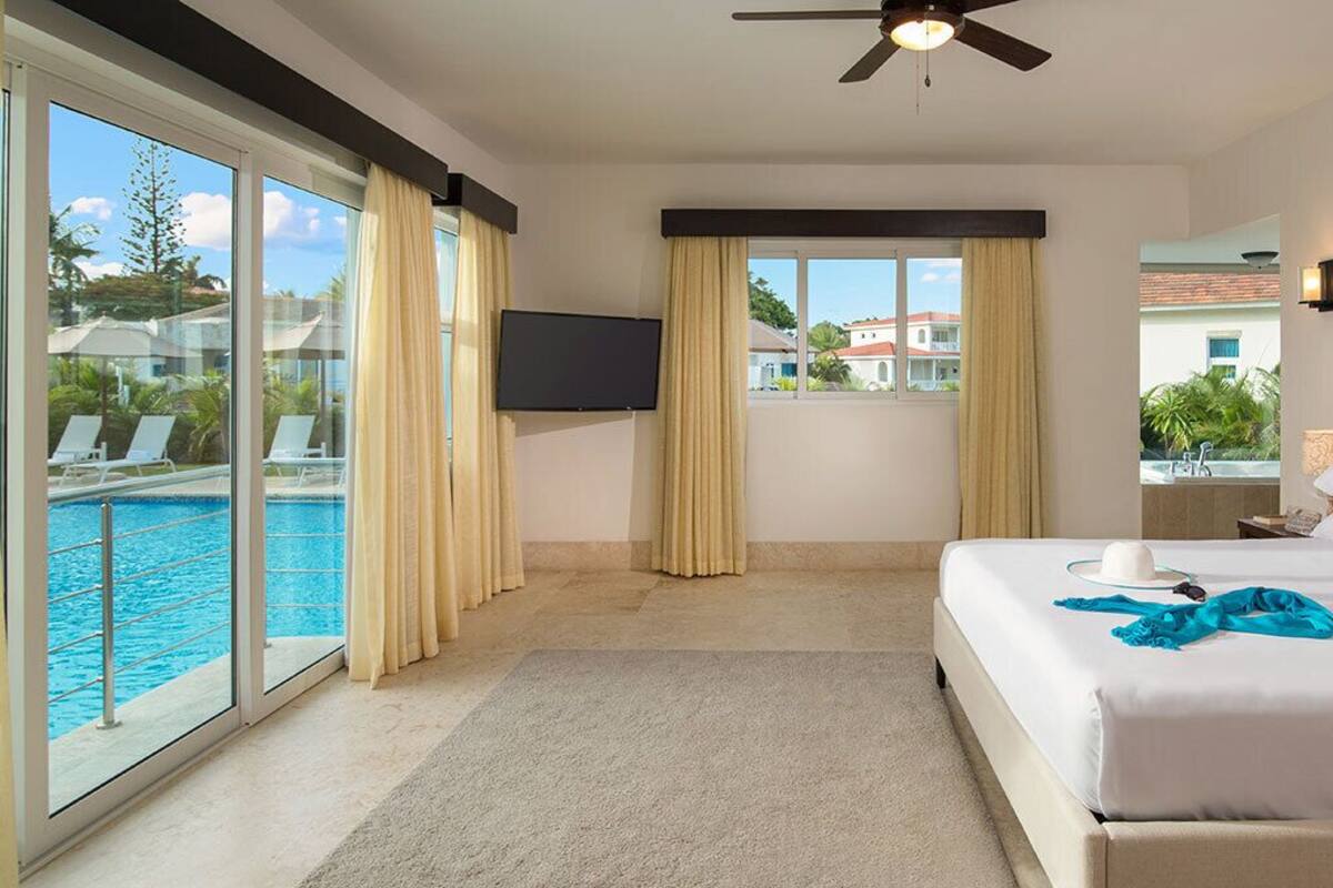 A hotel room with a large bed, a ceiling fan, a TV, a view of a swimming pool, and a balcony with a view of buildings.