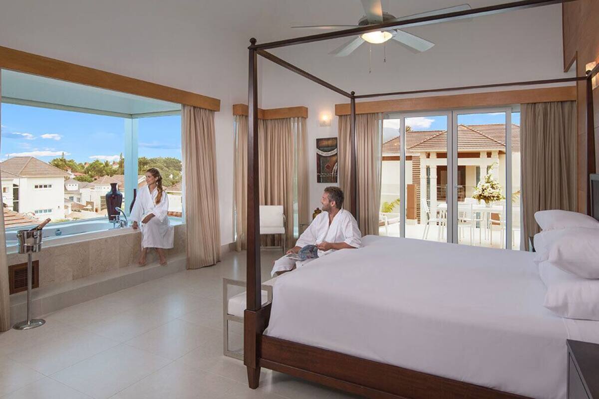 A spacious hotel room with a large bed, a balcony with a view of the surrounding area, and a ceiling fan.