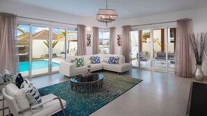 A spacious living room with a white sofa, a round coffee table, and a large window offering a view of a pool and outdoor seating. - Luxury Villas - Mandatory All-Inclusive (Puerto Plata)