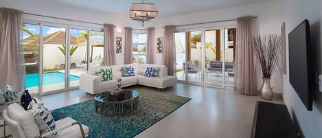 A spacious living room with a white sofa, a round coffee table, and a large window offering a view of a pool and outdoor seating.