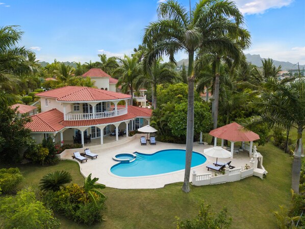 Una casa grande con piscina, rodeada de palmeras y montañas al fondo.