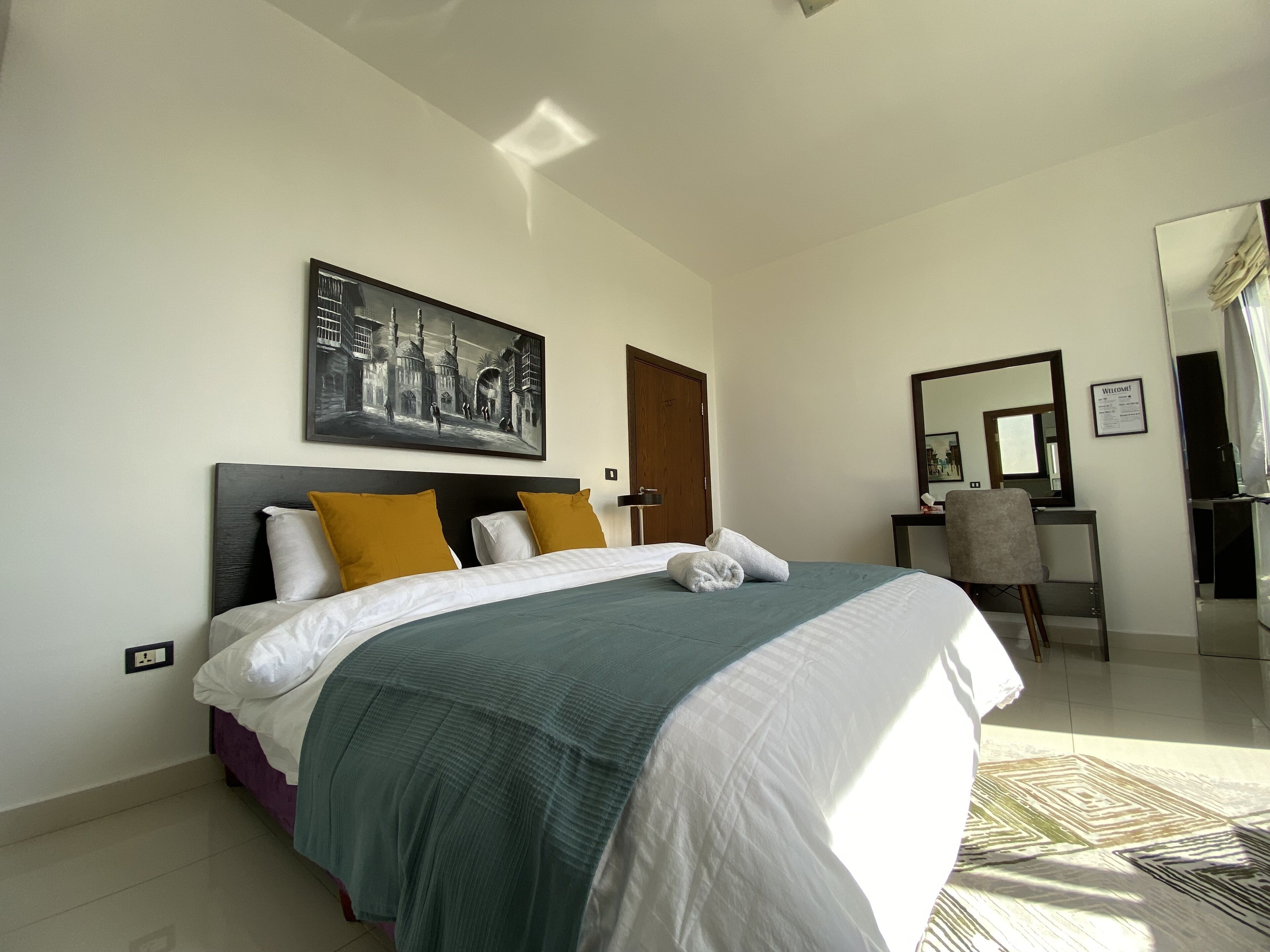 Quarto de hotel moderno com uma cama grande, um quadro na parede, uma escrivaninha com cadeira e um espelho.