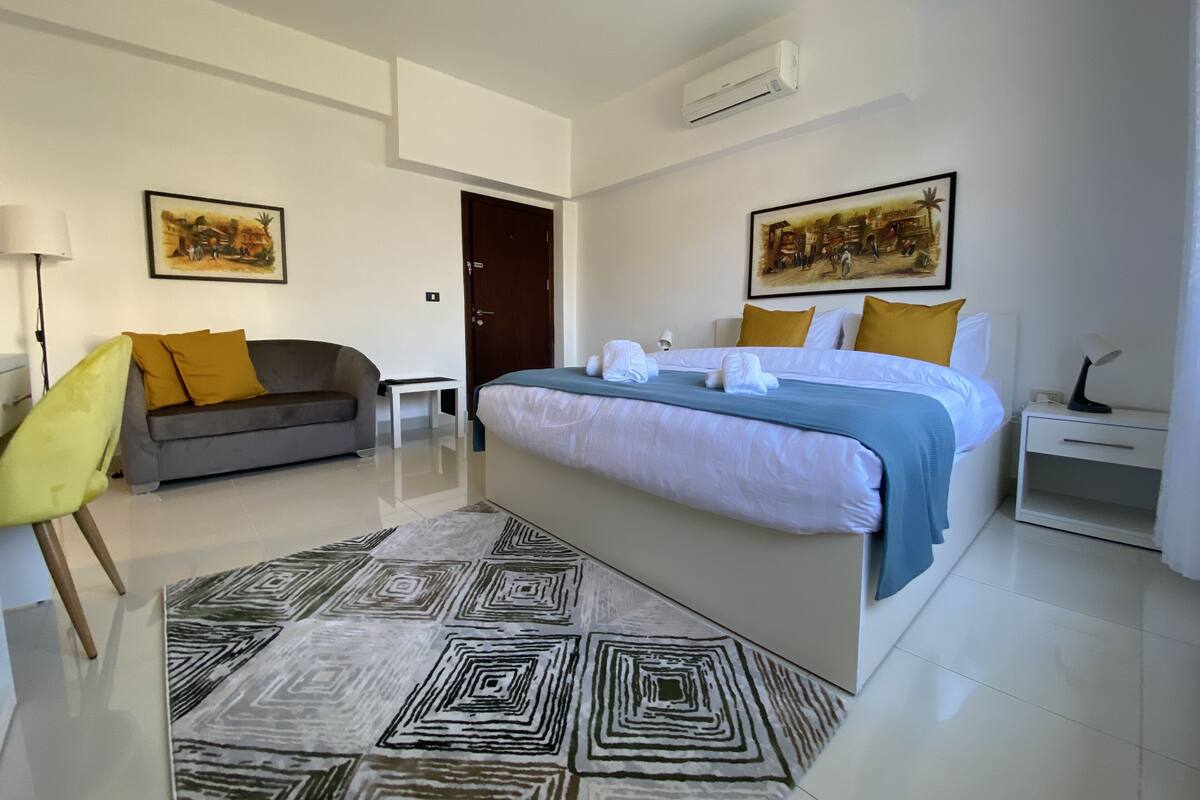 A modern hotel room with a large bed, a sofa, a small table, and a chair. There are two paintings on the walls and an air conditioning unit above the bed.