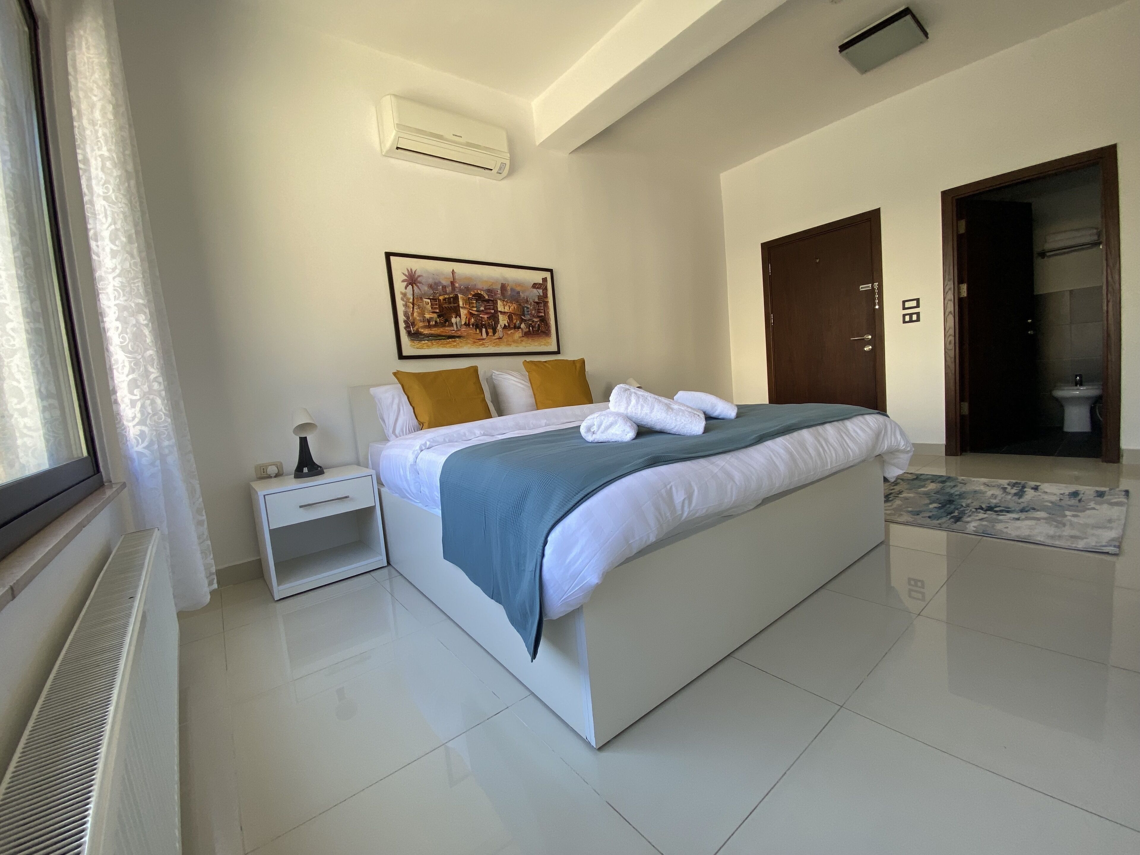 Una camera d'albergo moderna con un letto grande, un comodino con una lampada, un quadro appeso al muro e un bagno visibile attraverso una porta aperta.