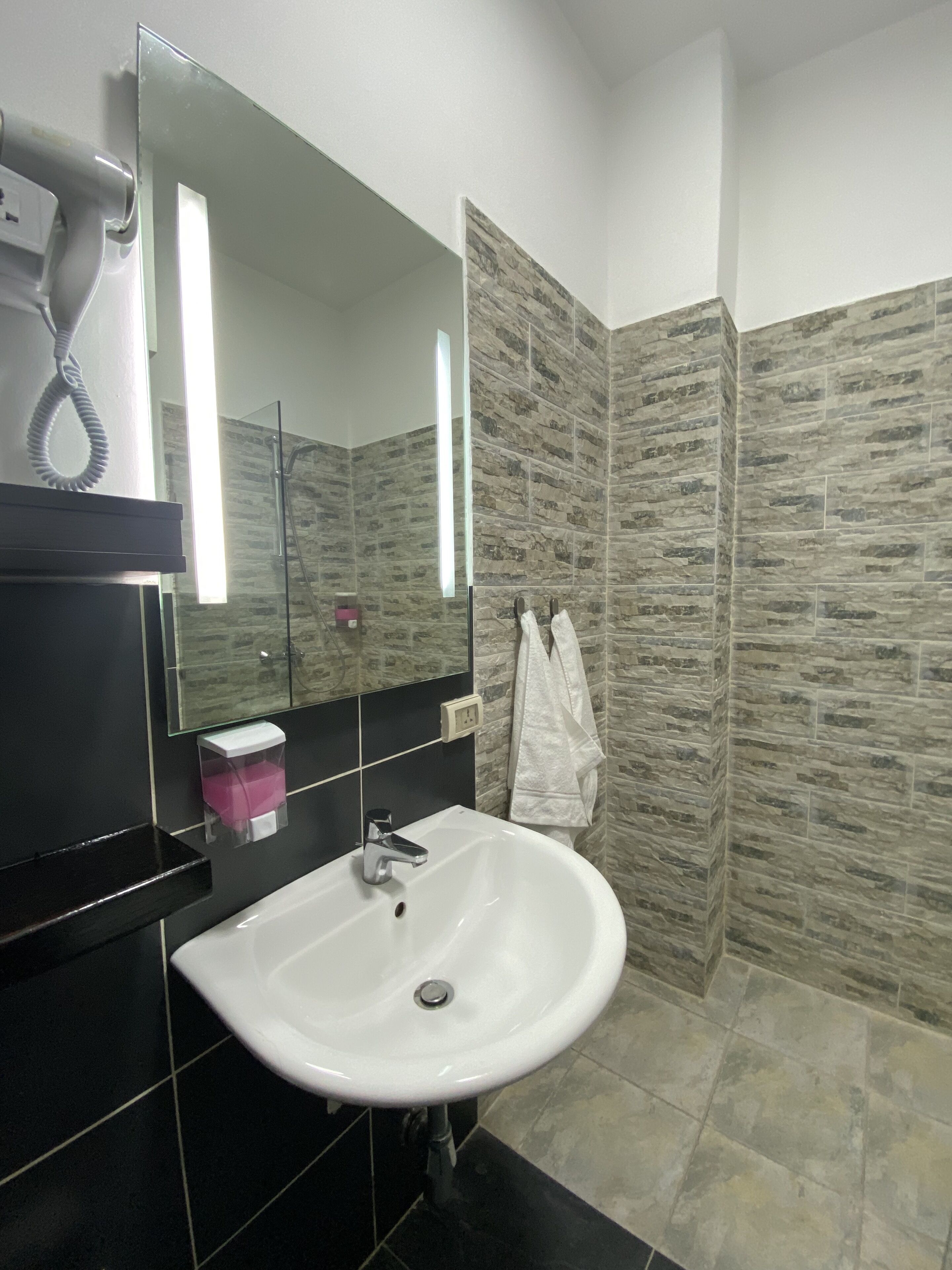Ein Badezimmer mit einem weißen Waschbecken, einem Spiegel, einem an der Wand hängenden Handtuch und einer Duschkabine mit Fliesen im Steinoptik.