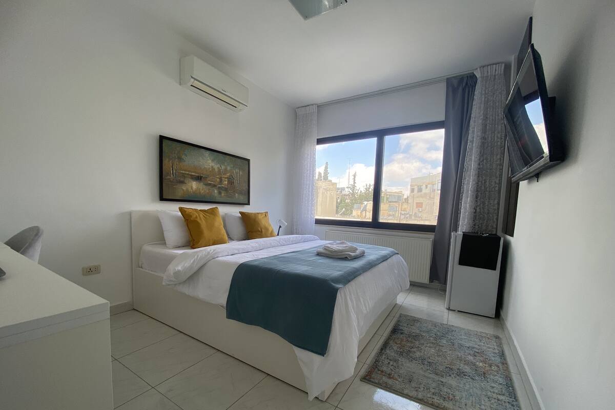 A hotel room with a bed, a desk, a TV mounted on the wall, and a window with a city view.