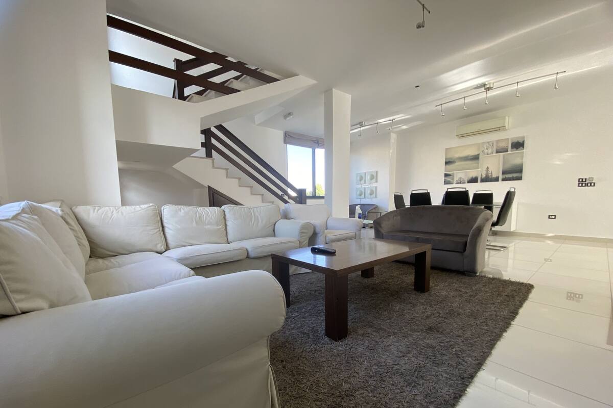 A modern living room with a sectional sofa, a coffee table, and a gray armchair. A staircase with wooden beams leads to an upper level.