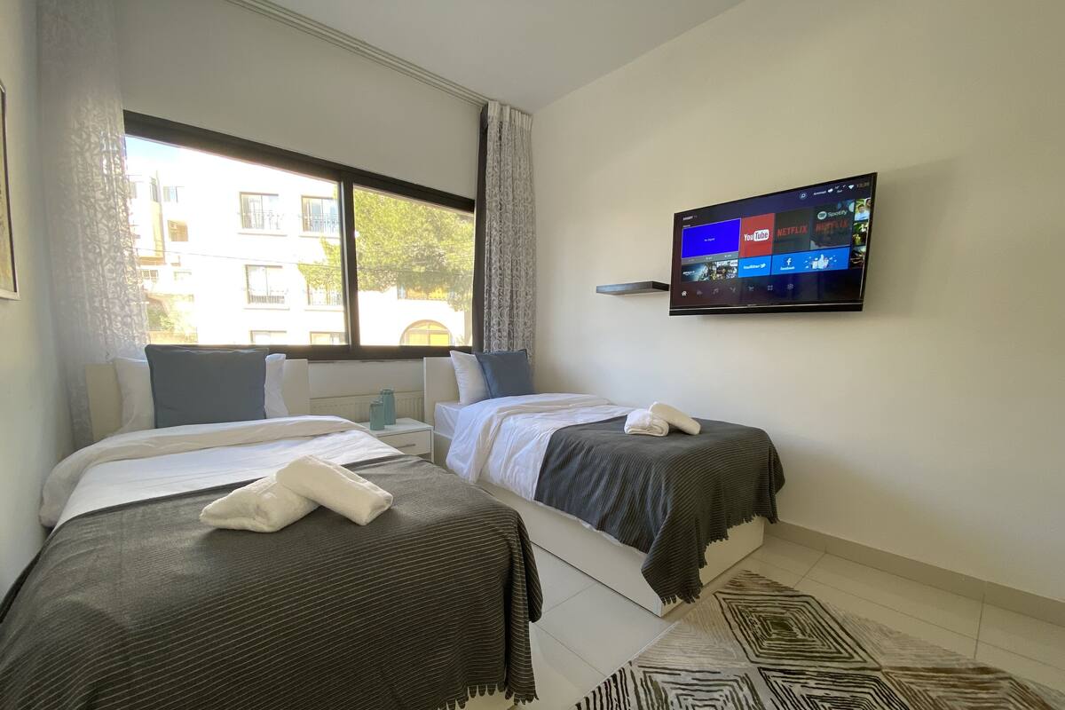 A hotel room with two beds, a TV mounted on the wall, and a window with a view of buildings outside.