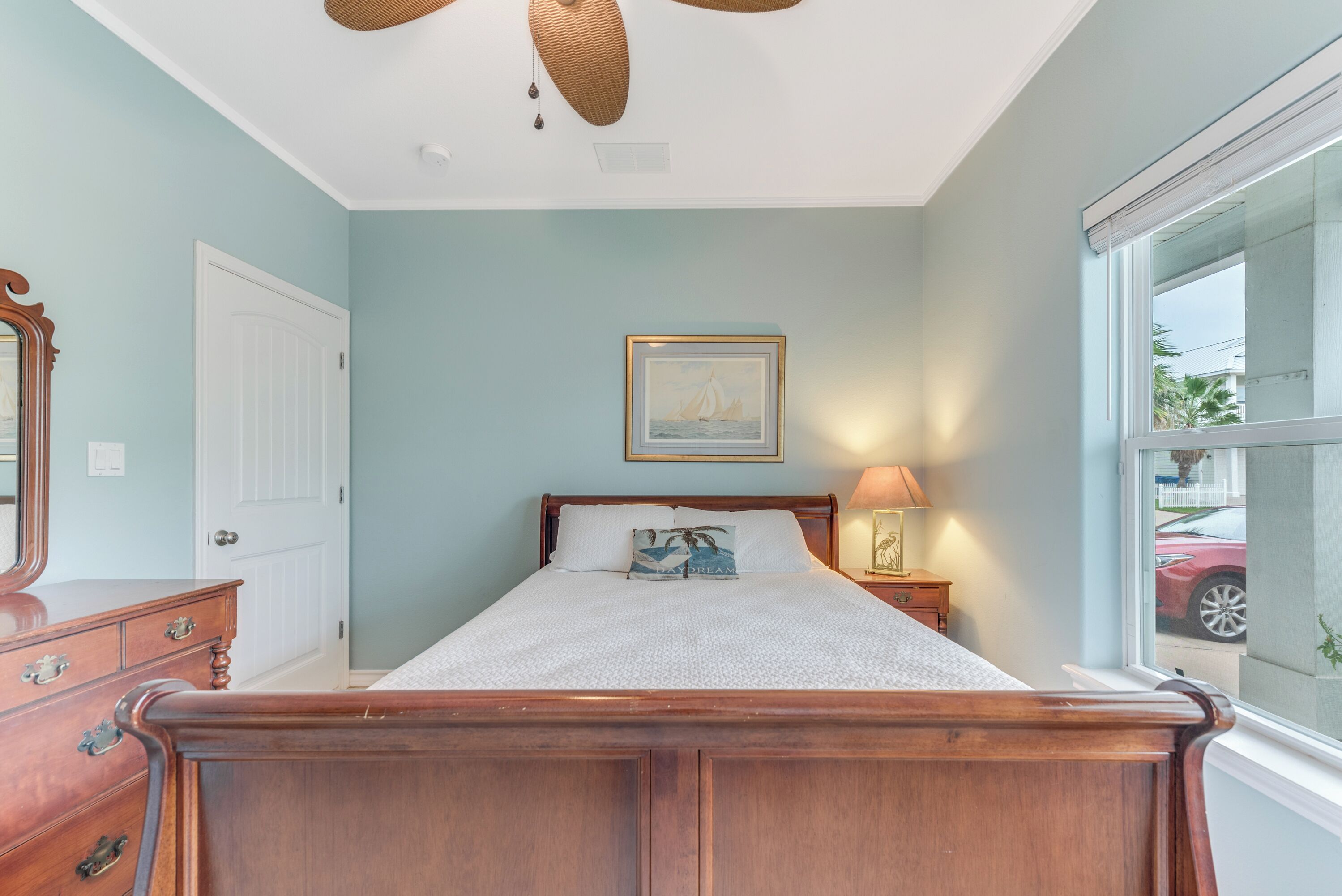 A bedroom with a wooden bed, a nightstand with a lamp, a framed picture on the wall, and a window with a view of the outside.