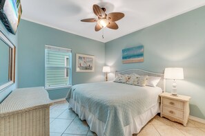 Un dormitorio con cama, mesita de noche, silla de mimbre, ventilador de techo, ventana con contraventanas y un cuadro en la pared.