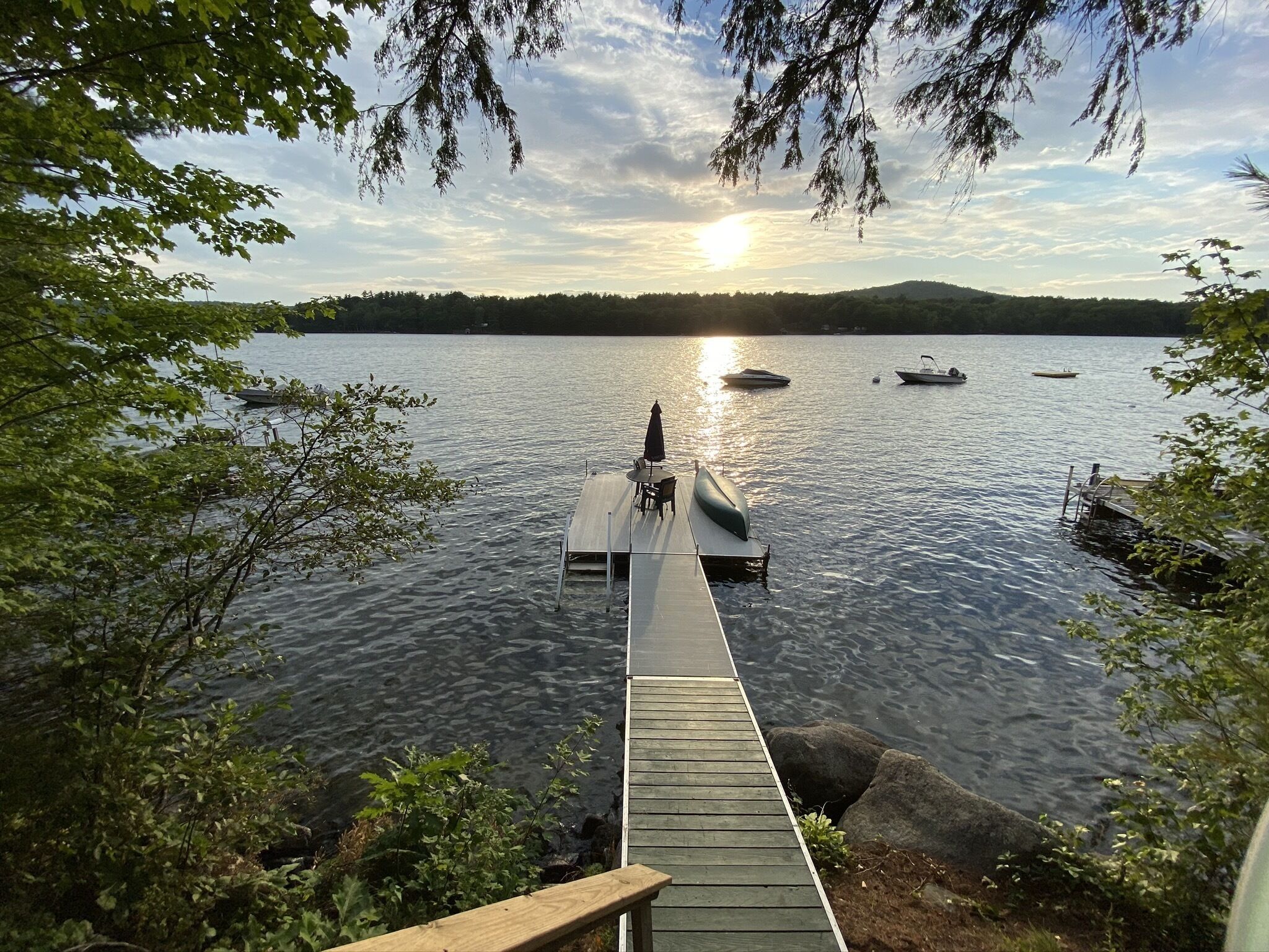 A wooden dock extending into a calm lake with boats, surrounded by trees and a distant mountain range.