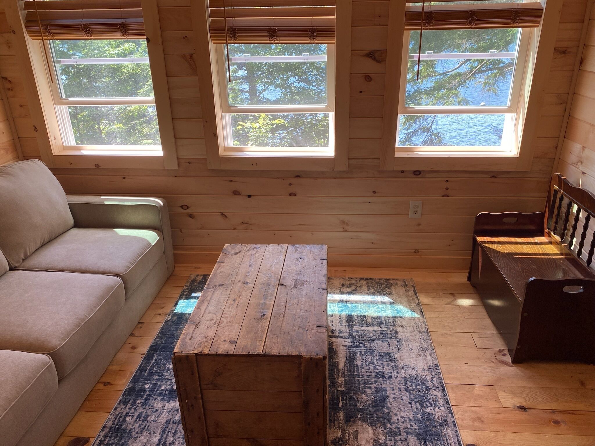 A cozy cabin interior with a sofa, a wooden table, and a bench. There are three windows providing natural light.