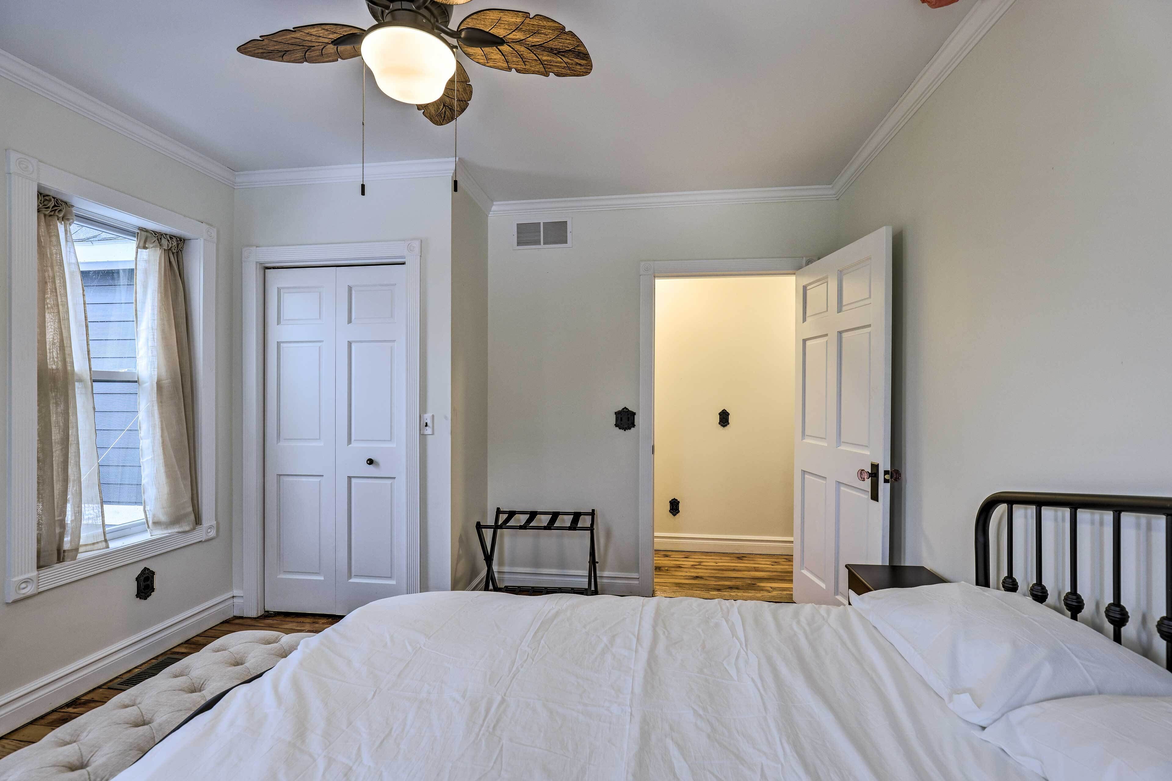 Un dormitorio con cama, ventilador de techo, ventana con cortinas y puerta que da a otra habitación.