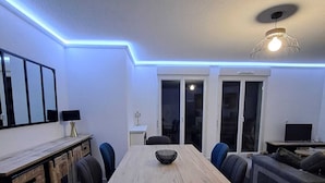 Un comedor moderno con una mesa de madera, sillas negras y un tazón en el centro. Se observa un sofá y un televisor al fondo.