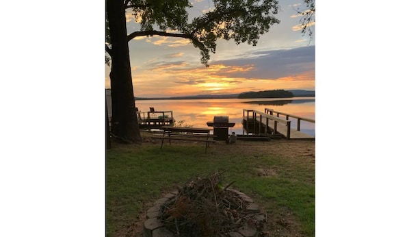 Une terrasse au bord du lac, avec un foyer, des bancs en bois et une vue sur le coucher de soleil sur l’eau.
