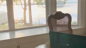 Une pièce dotée d’une grande fenêtre donnant sur un plan d’eau et des arbres. On aperçoit au premier plan une chaise en bois avec un dossier en osier.