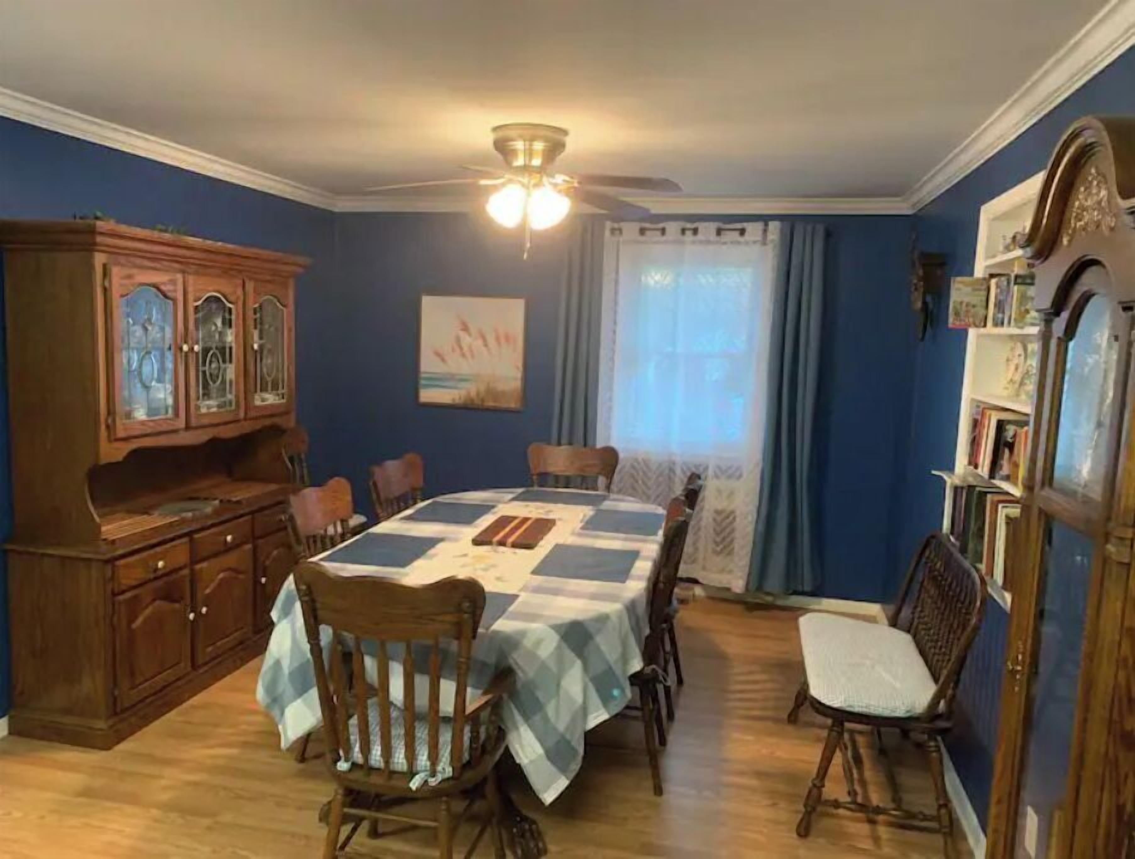 Une salle à manger avec une table dressée pour six personnes, des chaises en bois, un buffet aux portes vitrées, un ventilateur de plafond et une fenêtre avec des rideaux.