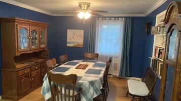 Une salle à manger avec une table dressée pour six personnes, des chaises en bois, un buffet aux portes vitrées, un ventilateur de plafond et une fenêtre avec des rideaux.