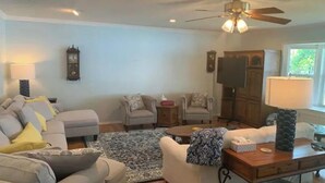 Un salon comprenant un canapé, des fauteuils, une table basse, un tapis, un bureau avec une lampe, un téléviseur et un ventilateur de plafond.