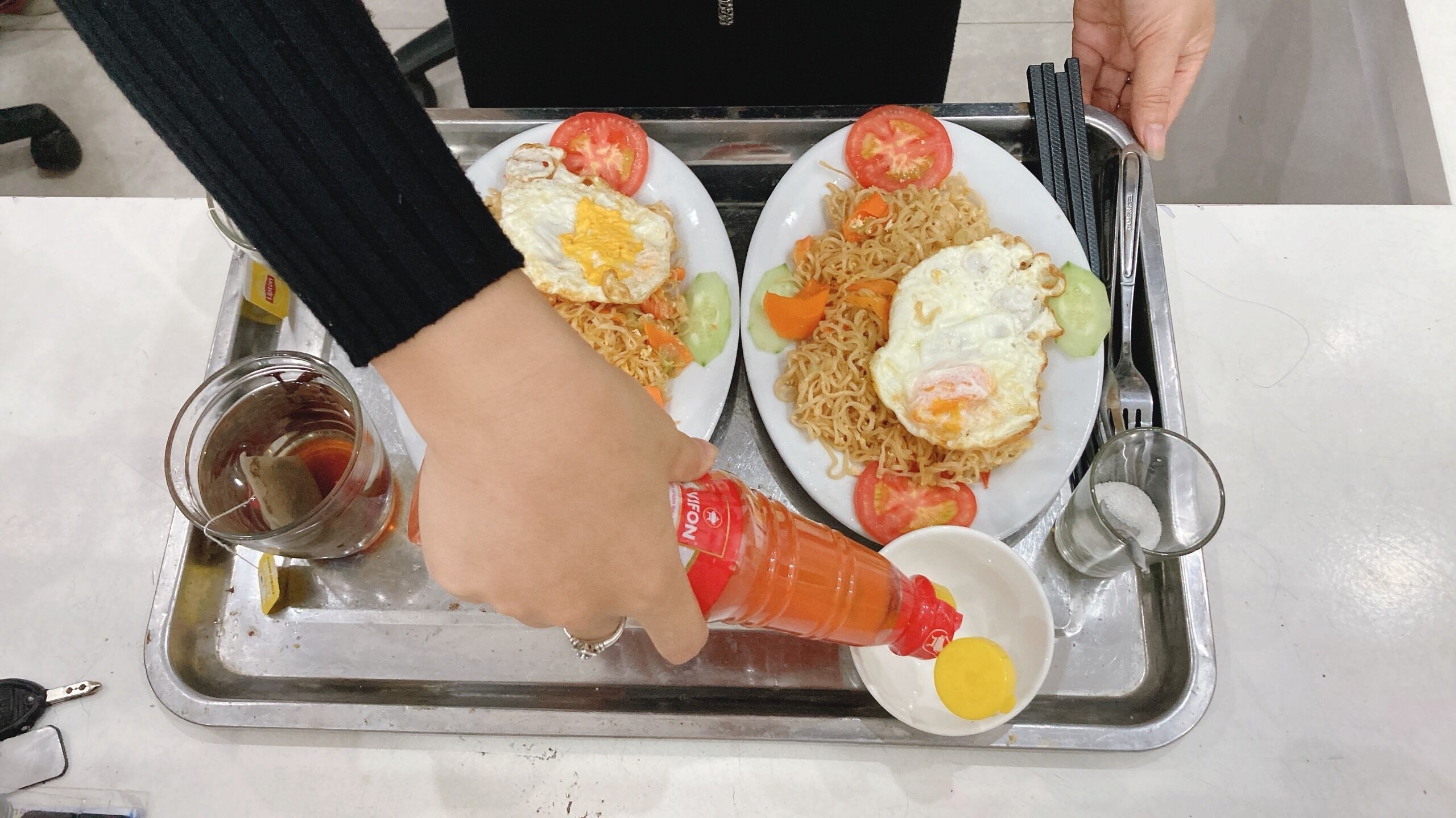 Un plateau avec deux assiettes de nourriture : l’une contient des nouilles et des œufs, et l’autre, des nouilles et un œuf frit. Une main verse une sauce rouge dans un petit bol.
