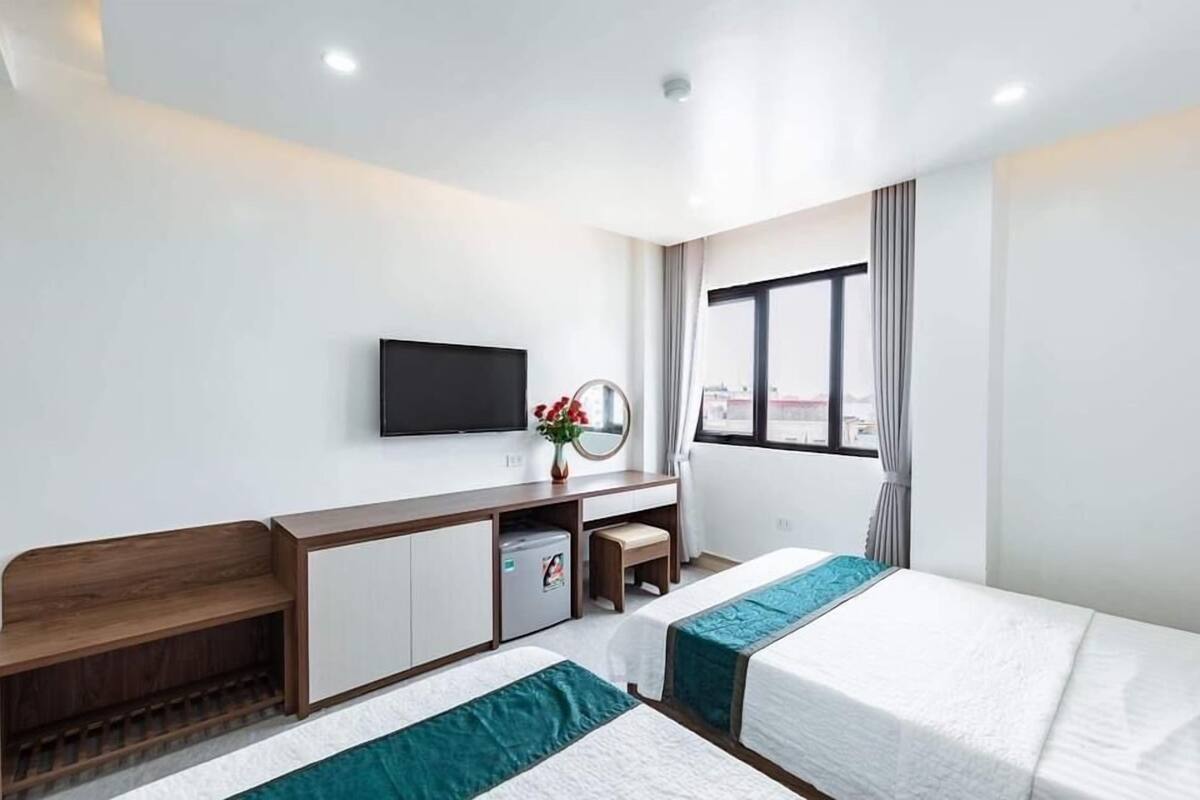 Habitación de hotel con dos camas, un escritorio con televisor, un refrigerador pequeño y una ventana con cortinas.