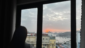 Eine Person steht an einem Fenster mit Blick auf eine Stadtlandschaft, darunter Gebäude und ein Gewässer, während die Sonne untergeht.