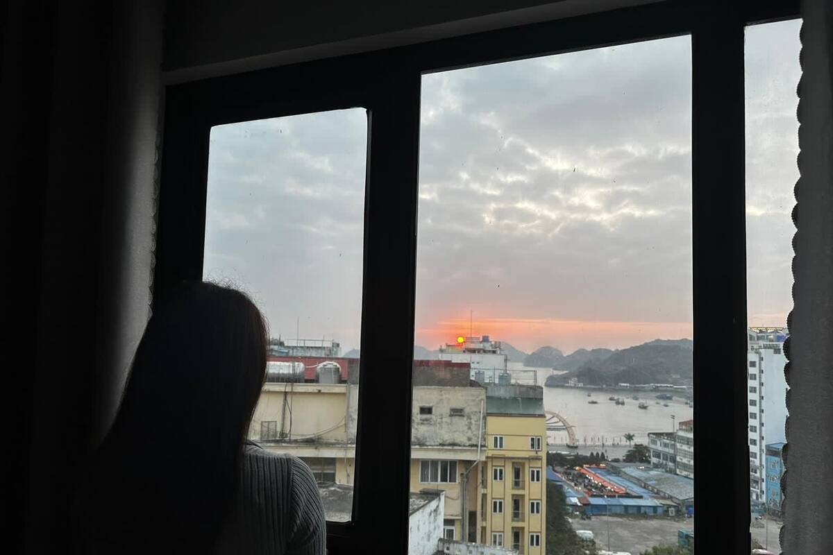 Una persona de pie junto a una ventana con vistas a un paisaje urbano que incluye edificios y una masa de agua al atardecer.