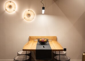 A dining area with a wooden table, two chairs, a black table runner, a bowl of fruit, and two wall-mounted light fixtures.