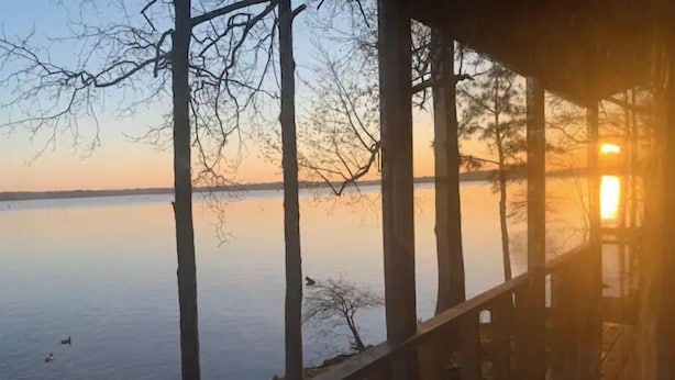 A wooden deck overlooking a calm lake with trees and a sunset in the background. - Lake house, amazing view, hot tub (Perryville)
