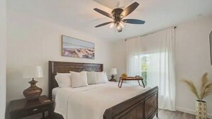 A bedroom with a bed, a nightstand, a lamp, a ceiling fan, a painting on the wall, and a window with curtains.