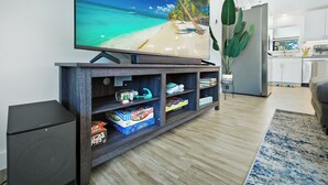 Olohuoneessa on suuri, litteänäyttöinen televisio puisen jalustan päällä, kirjahylly, jossa on monenlaisia esineitä, taustalla näkyy keittiönurkkaus ja huoneessa on myös suuri kasvi.
