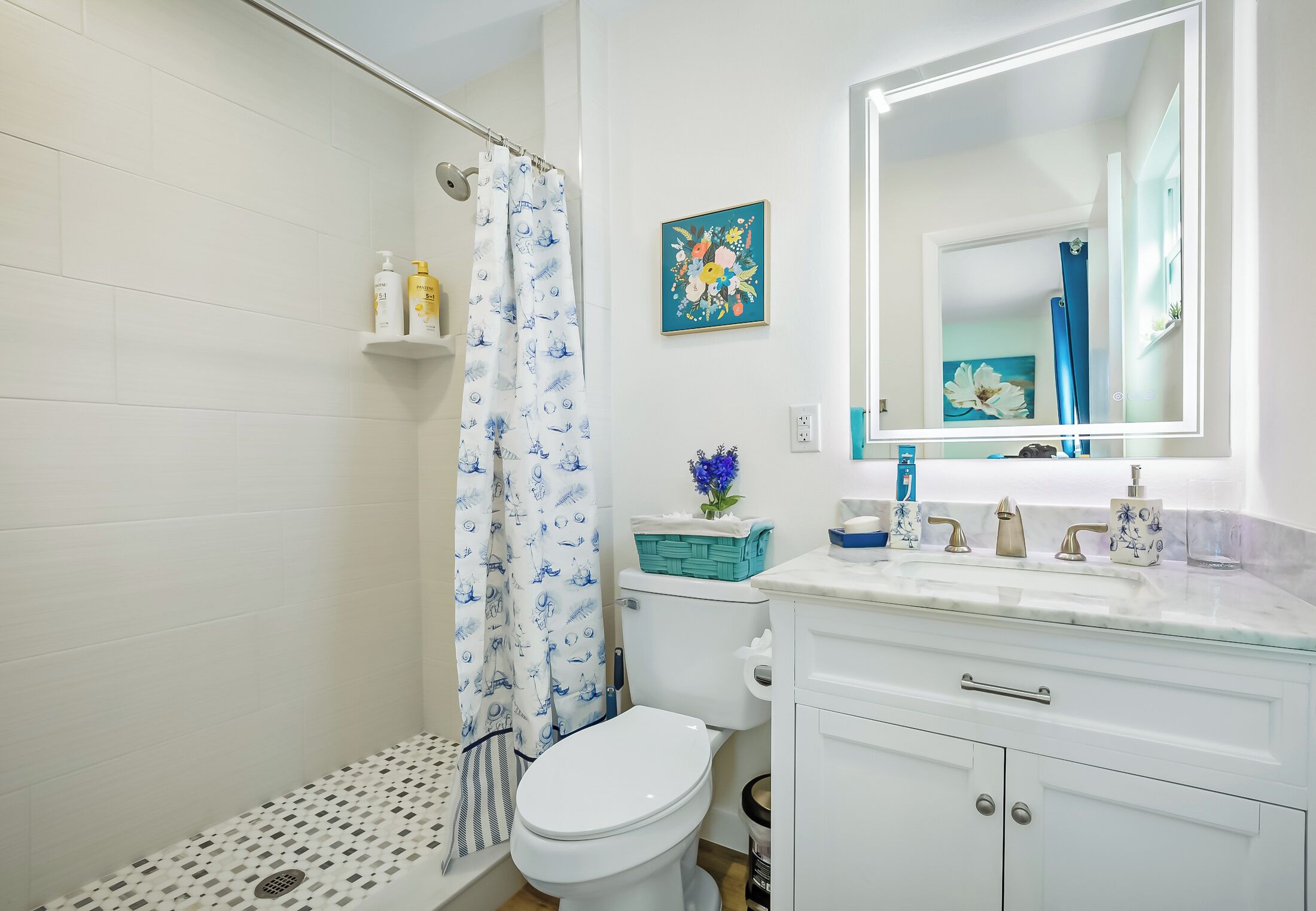 A bathroom with a white toilet, a shower with a blue and white patterned shower curtain, a white sink with a mirror above it, and a blue floral painting on the wall.