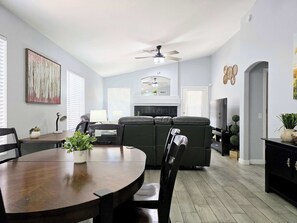 Een moderne woonkamer met een open haard, een eethoek met een houten tafel en een zithoek met een bank en stoelen.