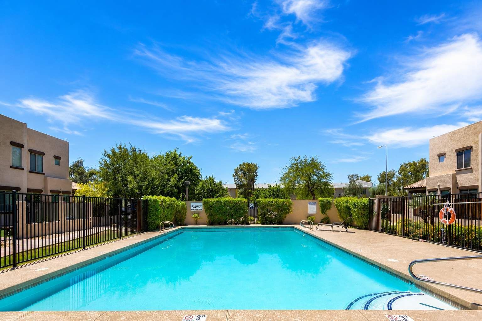 A rectangular swimming pool with a clear blue water surface, surrounded by a low fence, and set against a backdrop of buildings and trees under a clear sky.