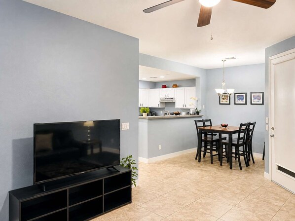 A living room with a flat-screen TV, a dining area with a table and chairs, and a kitchen in the background.
