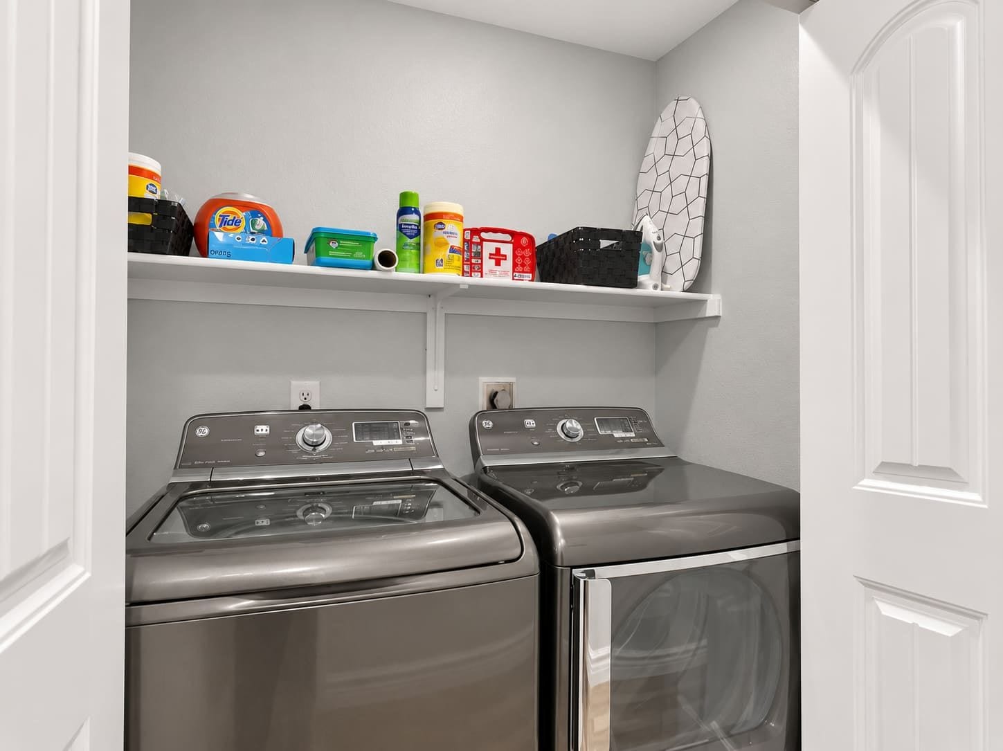 A laundry room with two front-loading washers and dryers, a shelf with various laundry supplies, and a surfboard.