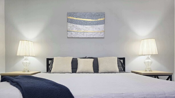 Una cama bien hecha con un edredón blanco y dos almohadas, flanqueada por dos lámparas de noche sobre mesitas de madera. Una manta azul está colocada sobre la cama, y una pequeña planta en maceta se encuentra sobre la mesita de noche a la derecha.