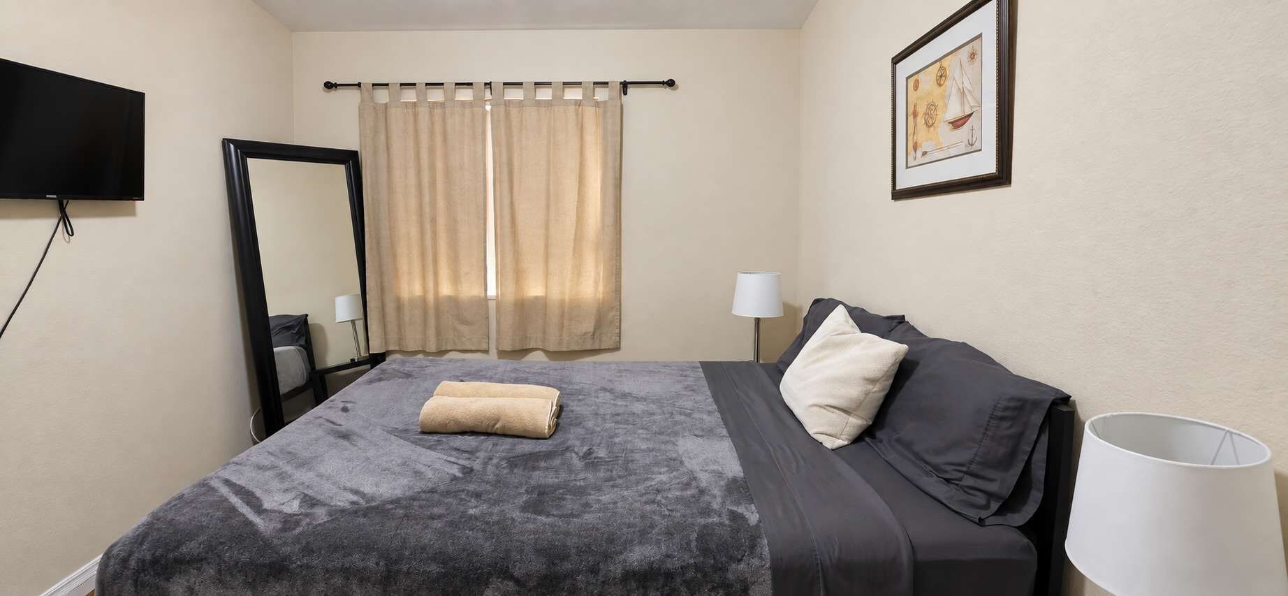 A hotel room with a bed, a flat-screen TV mounted on the wall, a window with curtains, a bedside lamp, and a framed picture on the wall.