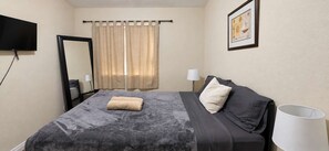A hotel room with a bed, a flat-screen TV mounted on the wall, a window with curtains, a bedside lamp, and a framed picture on the wall.