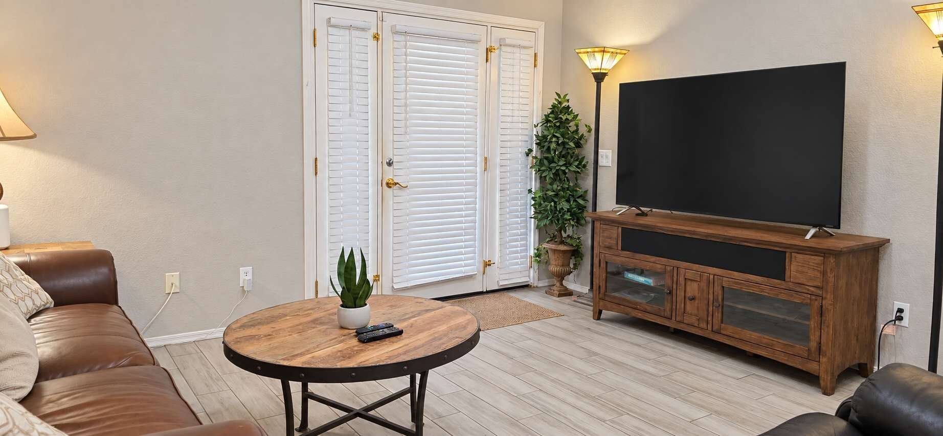 Un salon avec un canapé en cuir marron, une table basse ronde en bois agrémentée d’une plante, un téléviseur posé sur un meuble en bois et une porte-fenêtre coulissante avec des volets blancs.