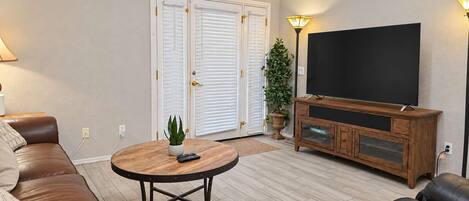 A living room with a brown leather sofa, a round wooden coffee table with a plant, a television on a wooden cabinet, and a sliding glass door with white shutters.